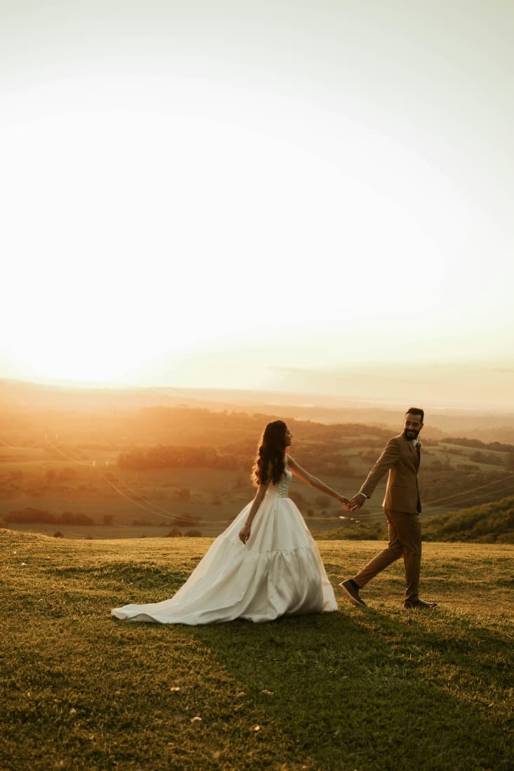A bride and groom holding hands while walking across a sunlit field