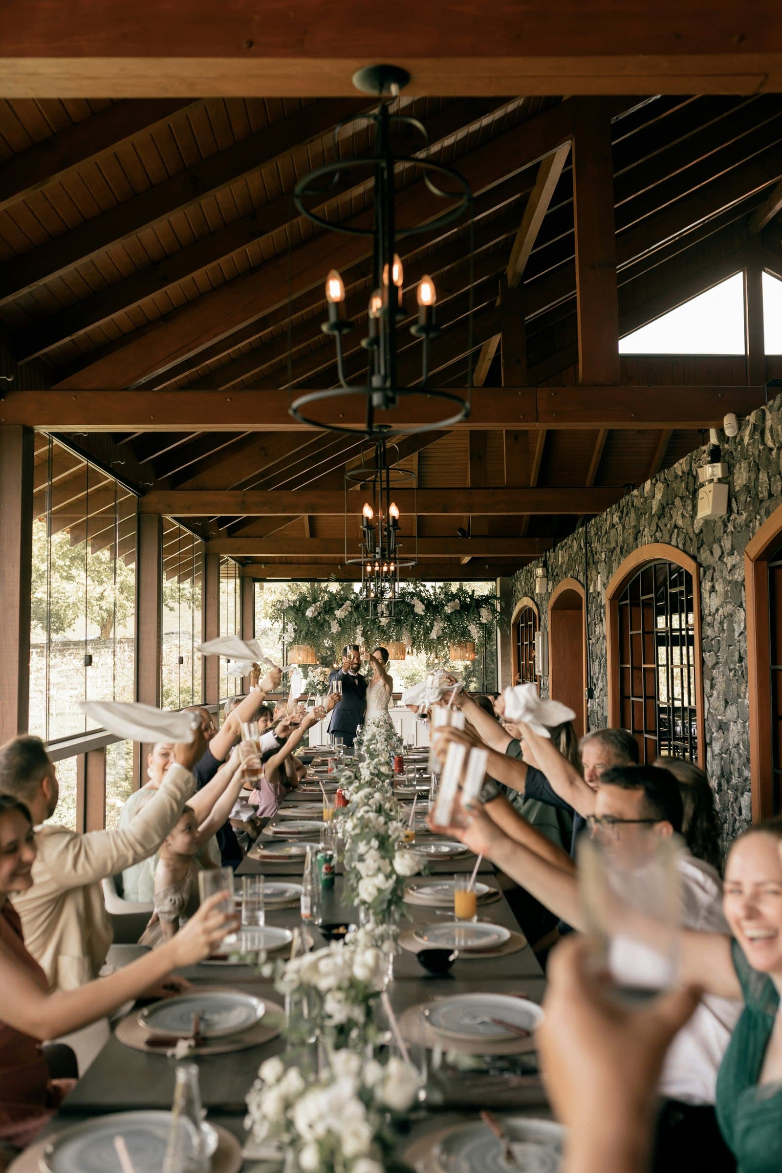 Wedding guests waving napkins around a long reception table decorated with white flowers