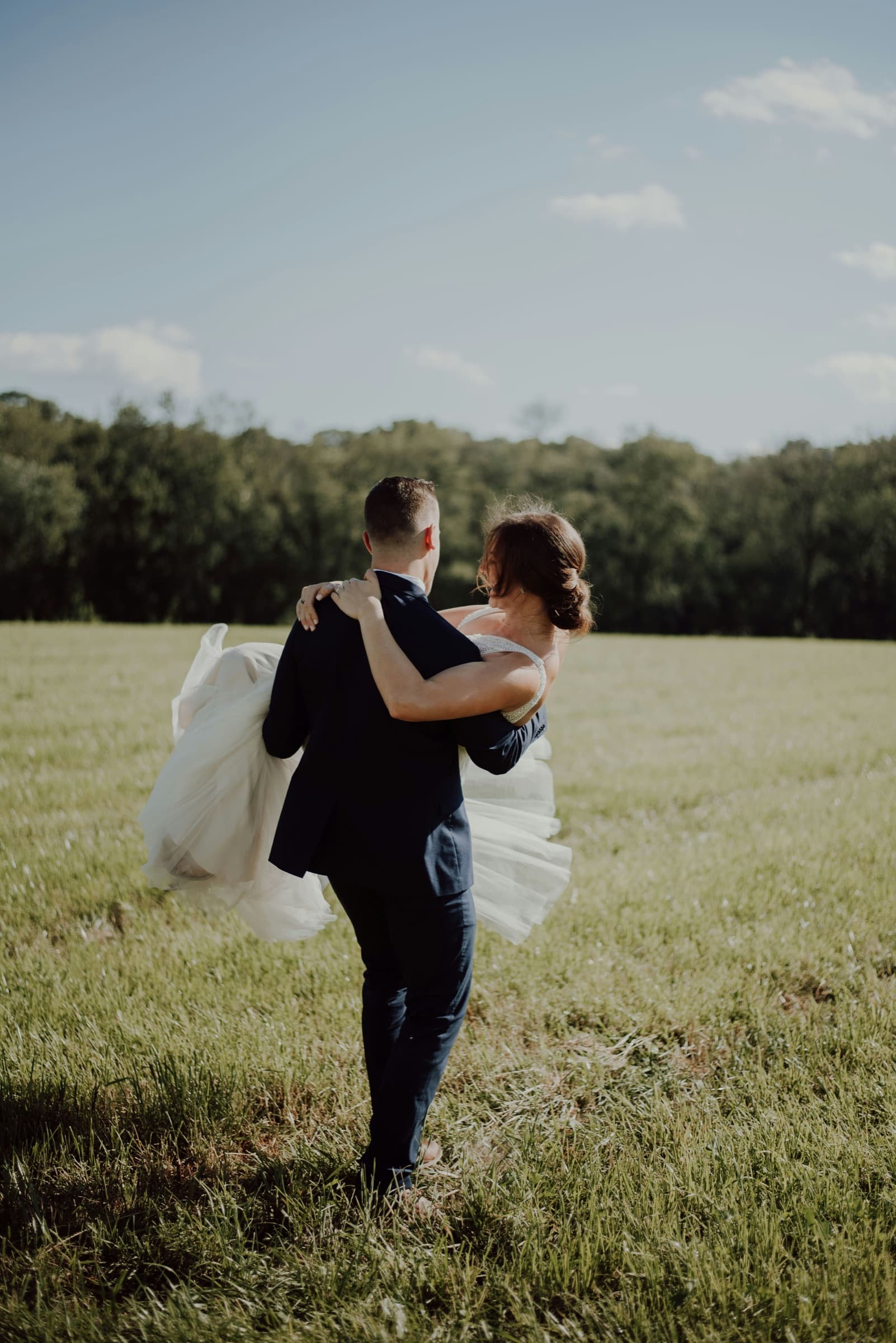 Groom carrying his bride across a sunlit grassy field on their wedding day